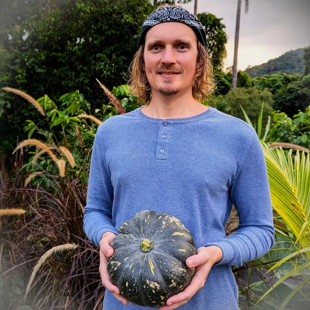 Person with blond hair holding a dark green pumpkin in a tropical garden with tall grasses and palm trees.